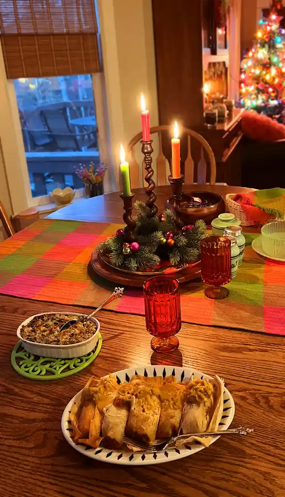 A cozy, warmly lit dining table is set for Christmas with a colorful plaid tablecloth, lit candles in wooden holders, and a…