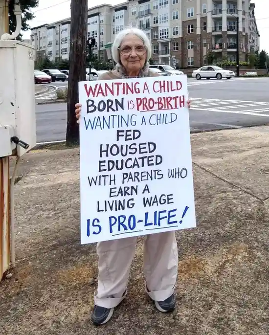 An elderly woman with white hair and glasses stands outdoors holding a handmade sign. The sign, written in red and blue…