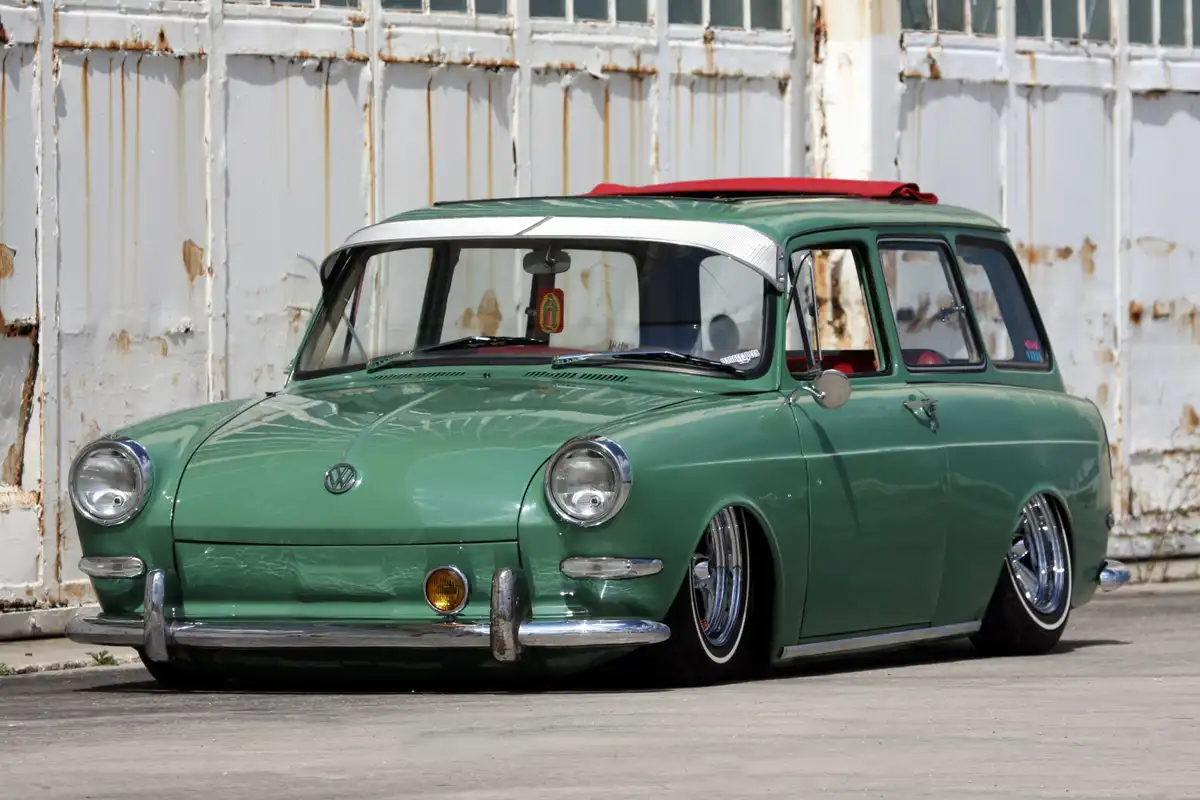 A green vintage Volkswagen station wagon with a red roof and chrome details is parked in front of a rusty white garage door…