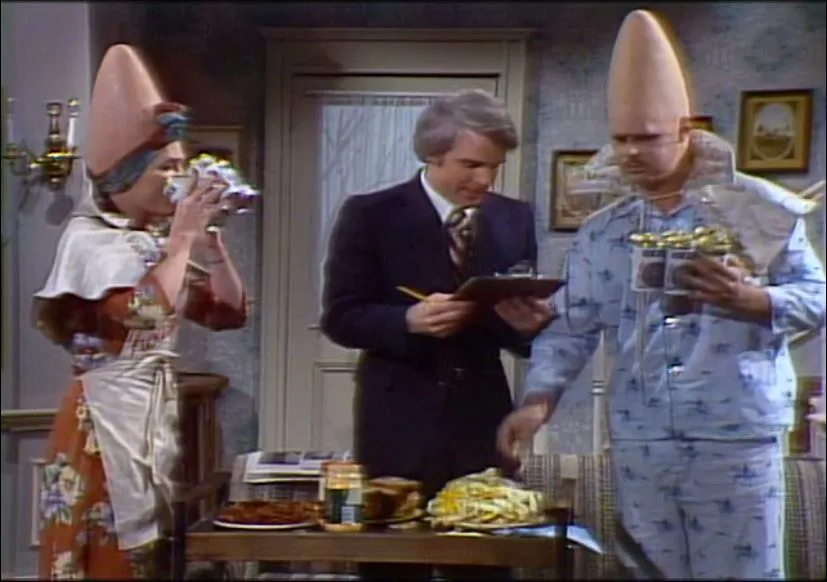In a scene from the TV show 'The Munsters,' two alien-like characters with cone-shaped heads stand beside a man in a suit…