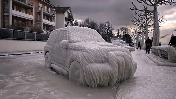 A car is completely encased in thick, textured ice and snow, resembling a snow sculpture, parked on a snowy street with…