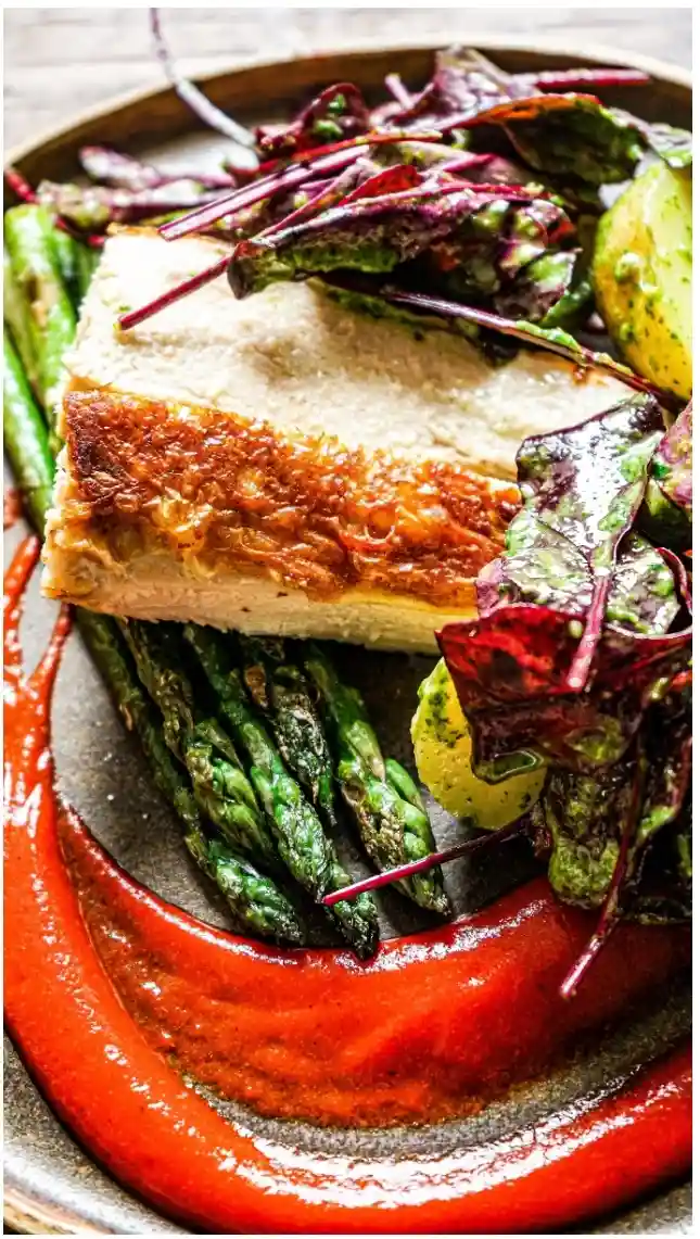 a close-up photo of a plated gourmet meal featuring a seared duck breast with a golden-brown crust, roasted green asparagus…