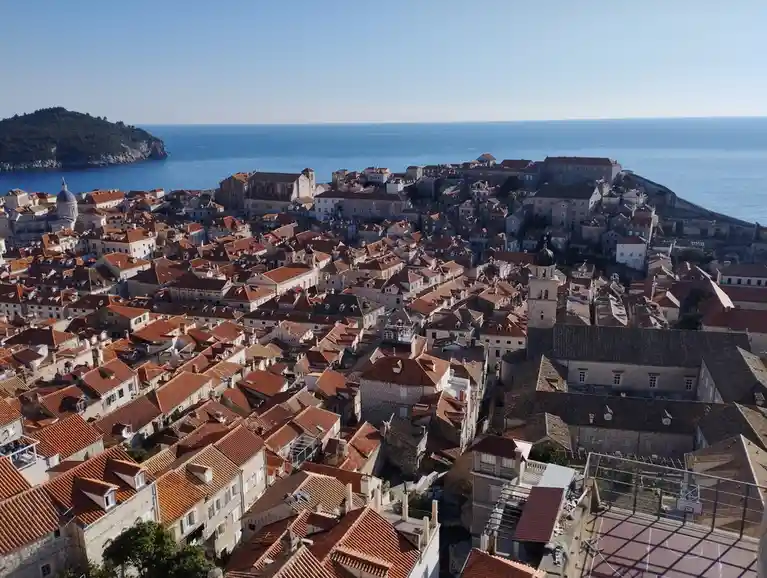 An aerial photo of a historic coastal town with red-tiled roofs, stone buildings, and a large ocean in the background. In…