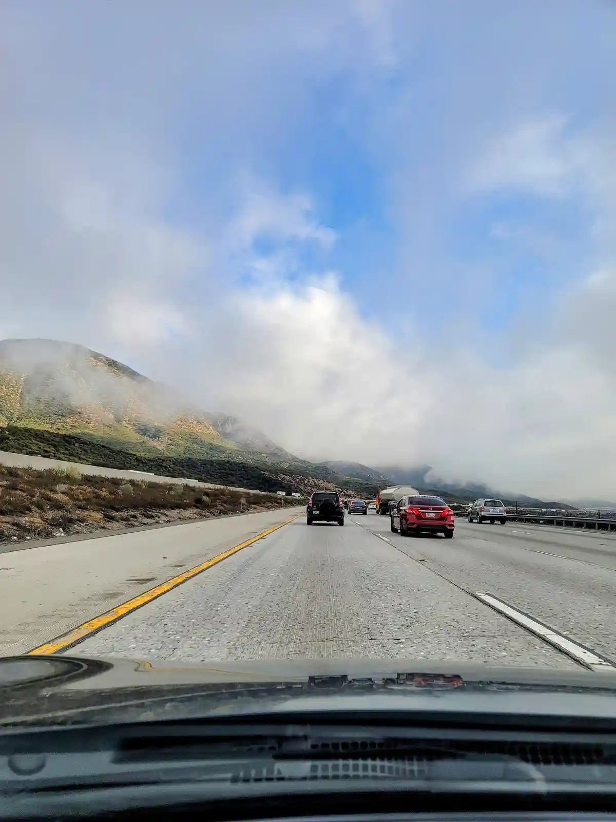 A point-of-view photo from inside a car on a multi-lane highway, showing several other vehicles ahead under a partly cloudy…