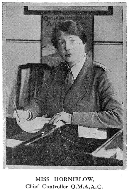 A vintage black-and-white photograph shows a woman in a dark, formal jacket with epaulets, seated at a desk and writing with…
