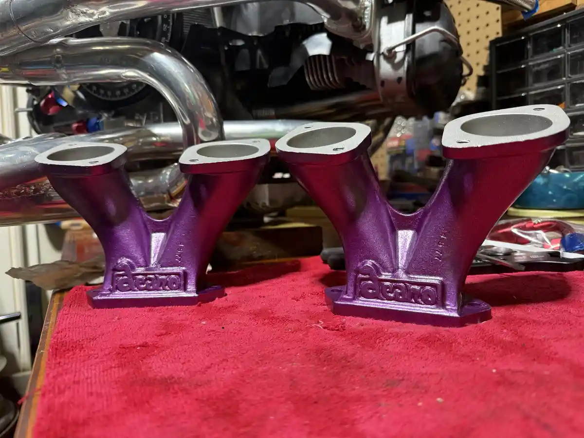 Two purple-painted aftermarket motorcycle intake manifolds with 'deano' embossed on them sit side-by-side on a red towel in…