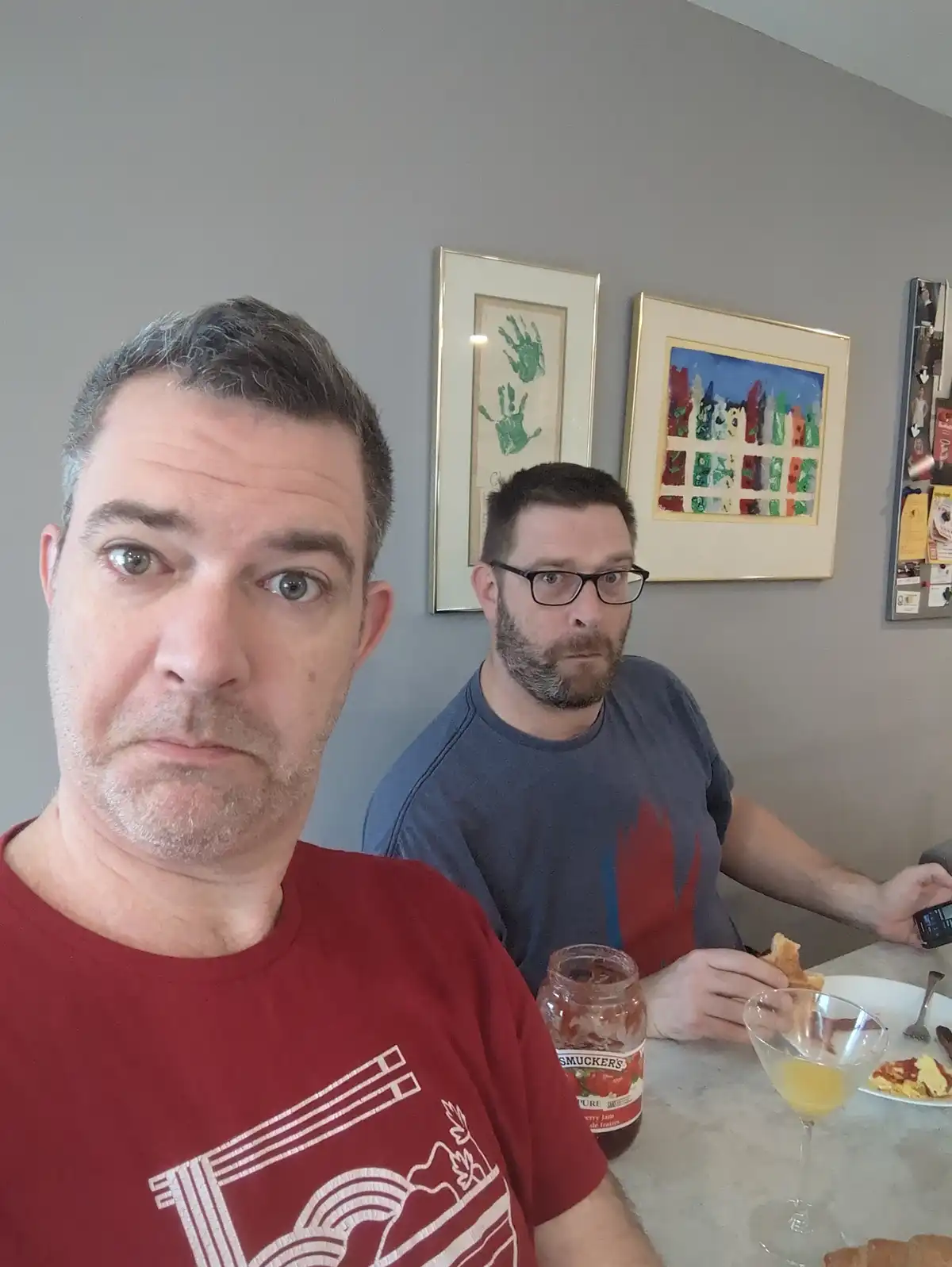 Two men sit at a breakfast table; the man in the foreground, wearing a red t-shirt, stares directly at the camera with a…