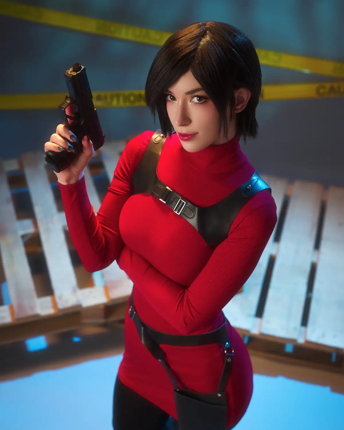 A woman with short dark hair, wearing a red turtleneck and black harness, holds a handgun while looking directly at the…