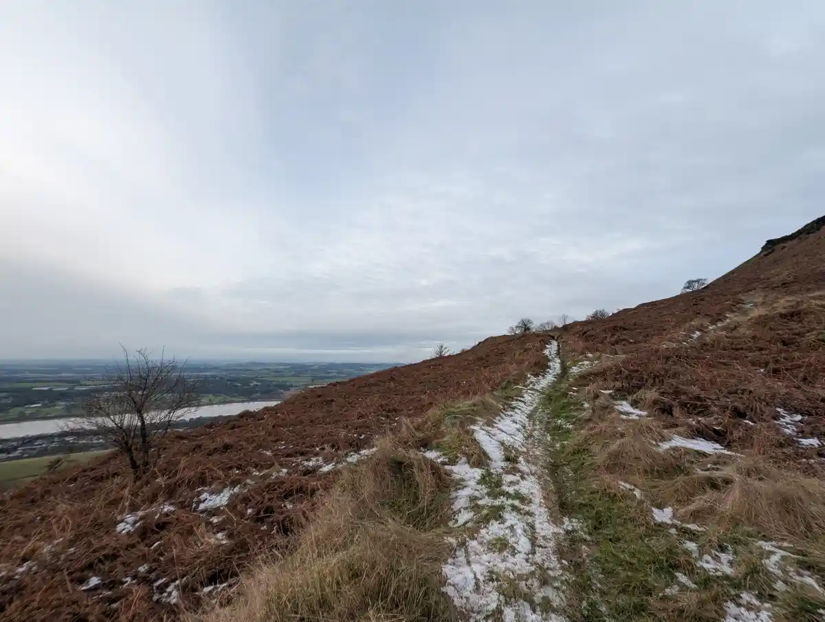 A narrow, snow-dusted hiking trail winds up a grassy, brown hillside under an overcast sky, with a distant valley and river…