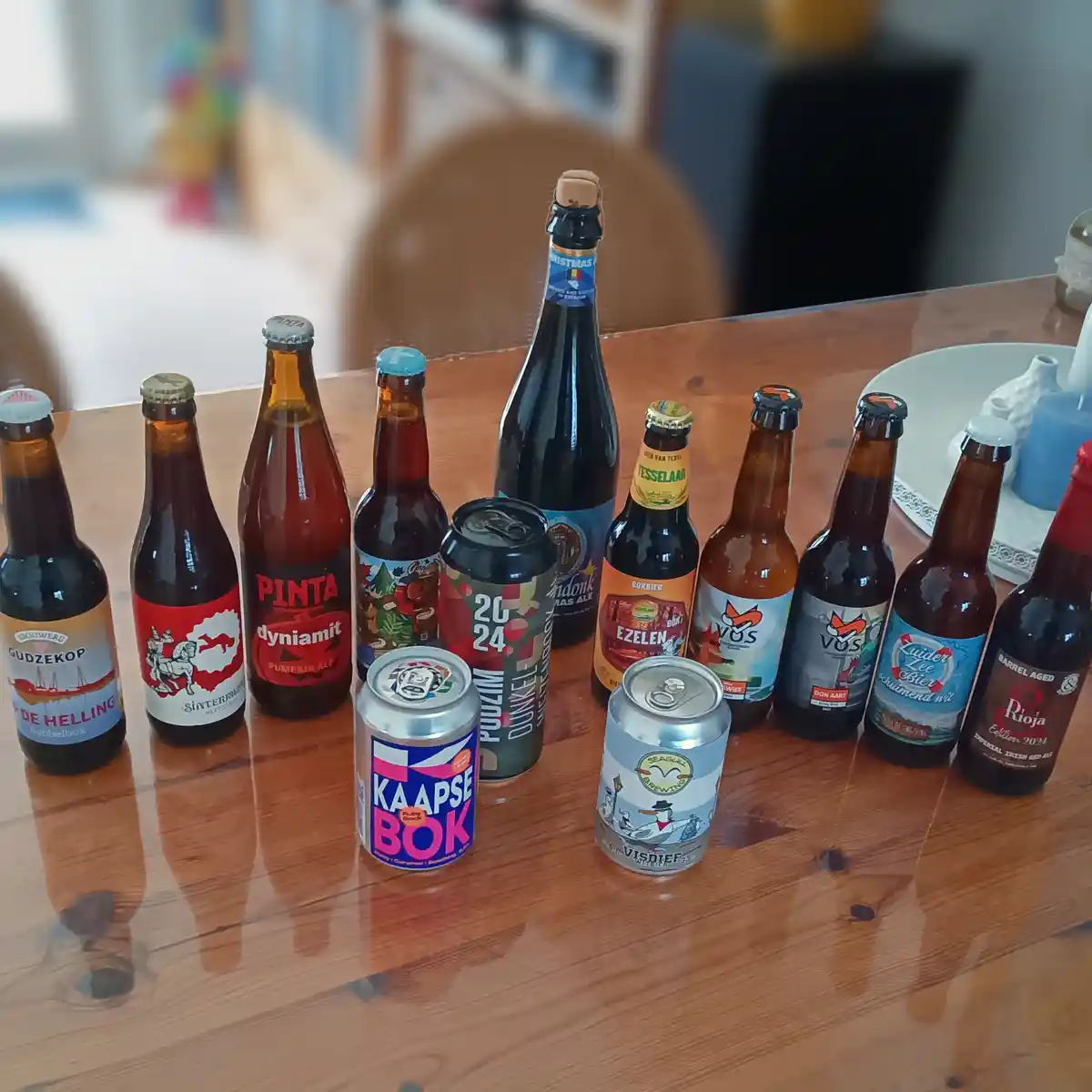 A wooden table displays a lineup of 12 different beer bottles and cans, including brands like Pinta, Kaapse Bok, and Vos…