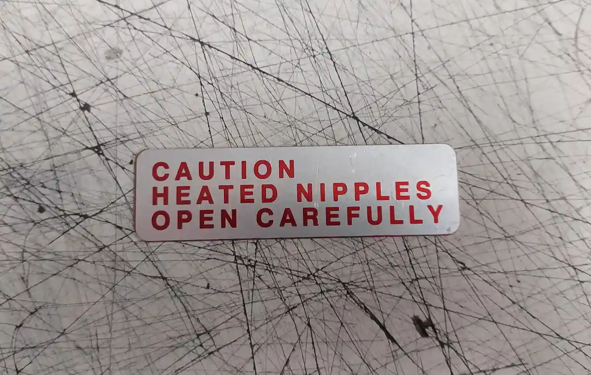 A close-up photo shows a rectangular white warning label with red uppercase text reading 'CAUTION HEATED NIPPLES OPEN…