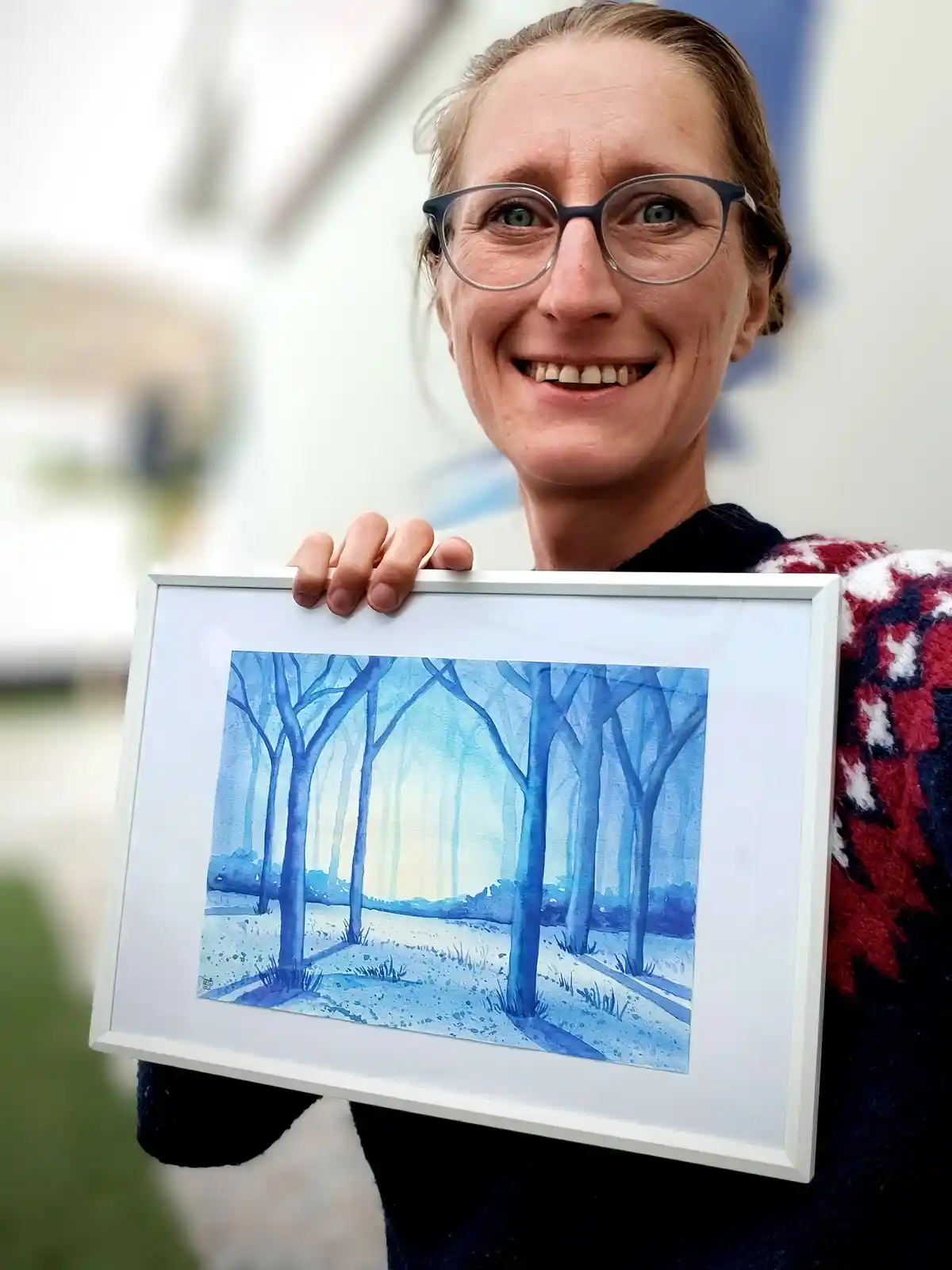 A woman with glasses and a red-and-white patterned sweater holds up a framed watercolor painting of a blue-toned forest…