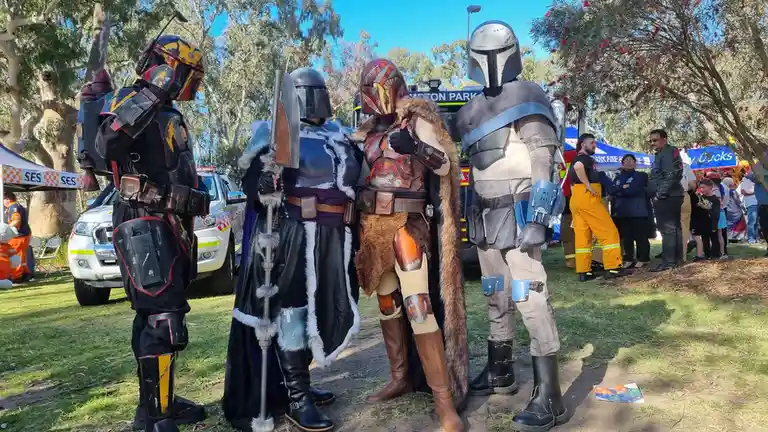 Four people in detailed Mandalorian armor costumes from Star Wars stand together at an outdoor event, with one giving a…