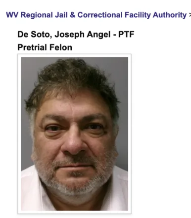 A mugshot of a middle-aged man with graying hair and a beard, wearing a white shirt, with official text above identifying…