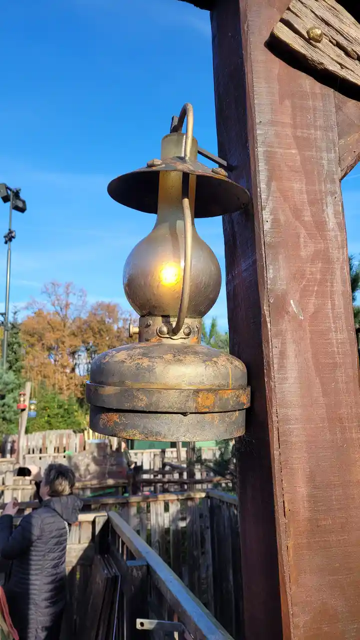 A close-up of a vintage-style, rusted metal lantern with a glowing bulb, mounted on a wooden post outdoors under a clear…