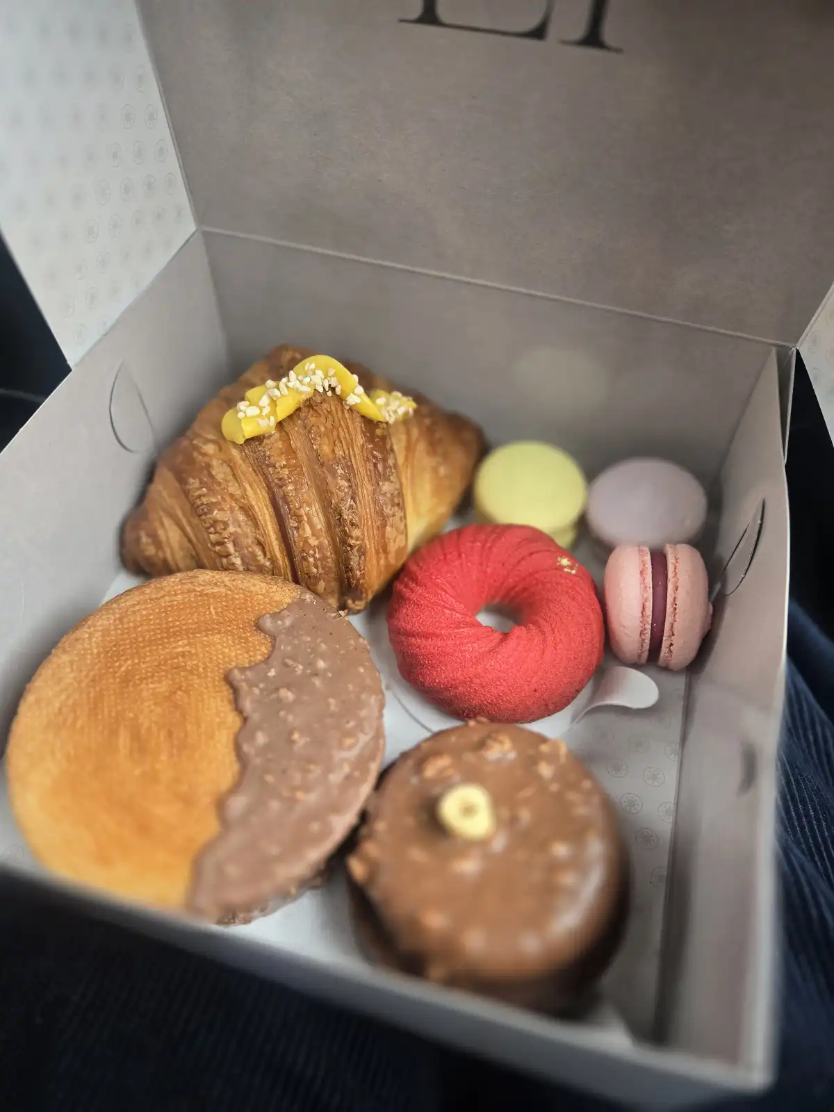 A close-up shot of an open gray bakery box containing assorted pastries: a croissant with yellow icing, colorful macarons, a…