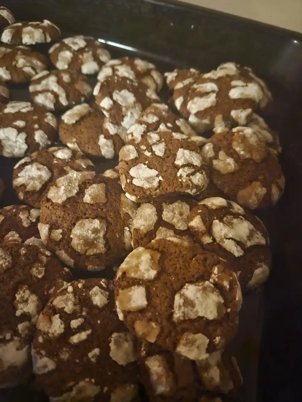 A close-up photo shows a dark baking tray filled with freshly baked chocolate crinkle cookies, each with a distinctive…
