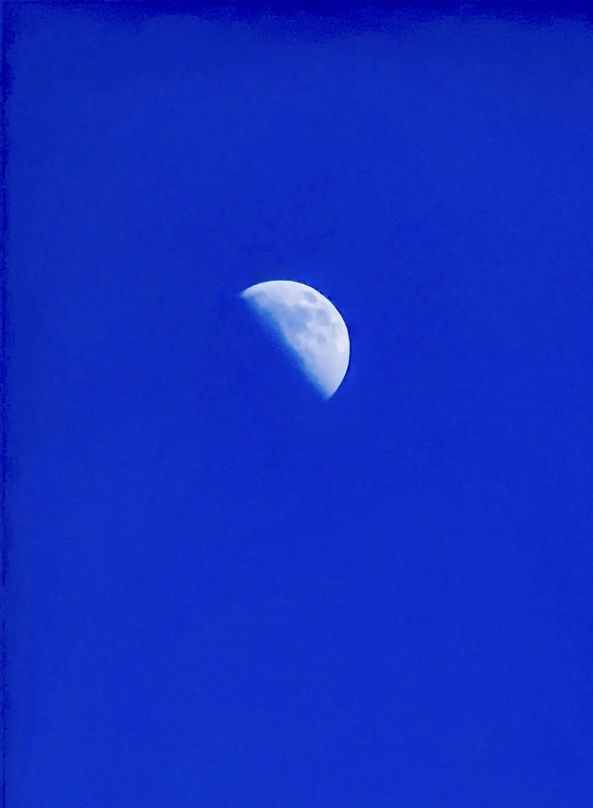 A crescent moon is visible against a solid, deep blue sky, with the moon's surface showing craters and shadows. The image…