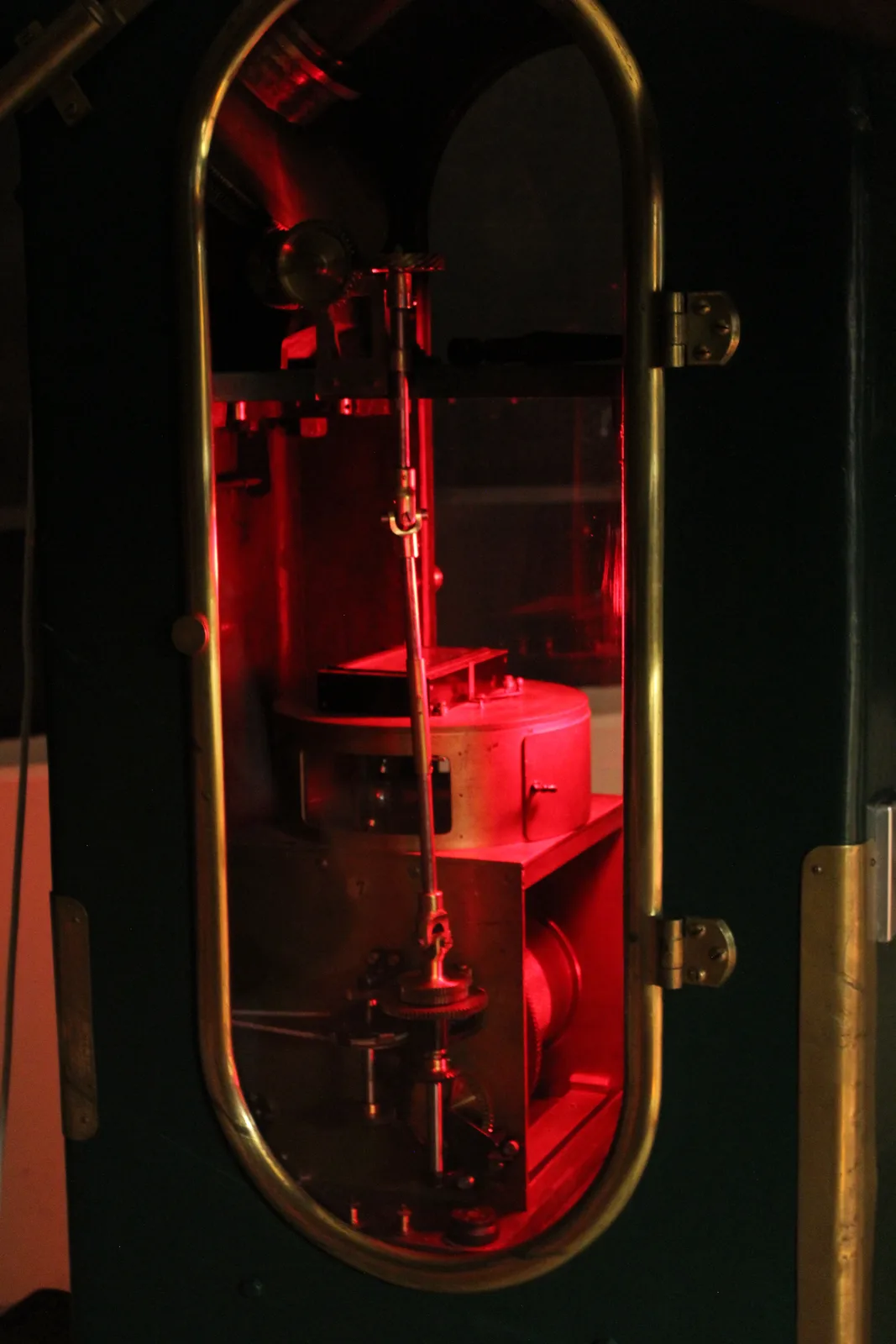 A close-up shot of a complex, vintage mechanical device inside a brass-framed enclosure, bathed in a red glow from within…