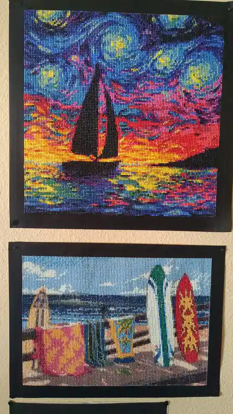 Two bead art pictures hang on a wall; the top one is a colorful sailboat scene, the bottom one shows surfboards and beach…