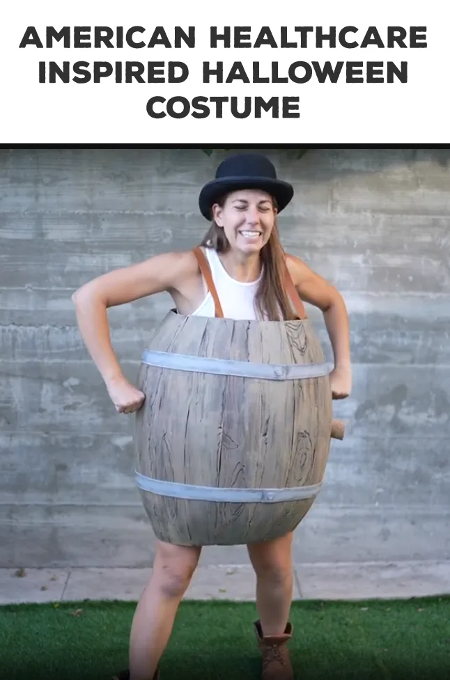 A woman wearing a large wooden barrel costume with metal bands, a white tank top, and a black bowler hat, standing with…