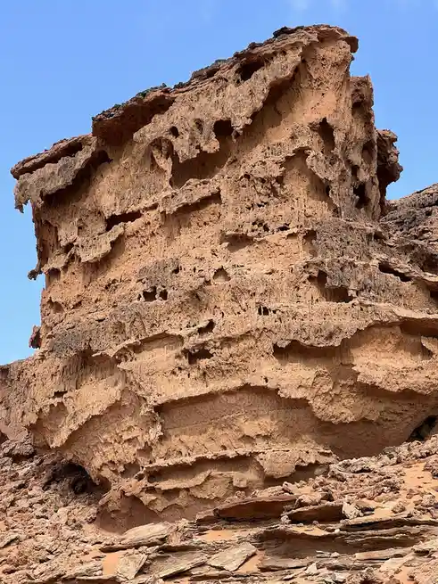 A tall, weathered, reddish-brown rock formation with numerous holes and layered erosion patterns stands against a clear blue…