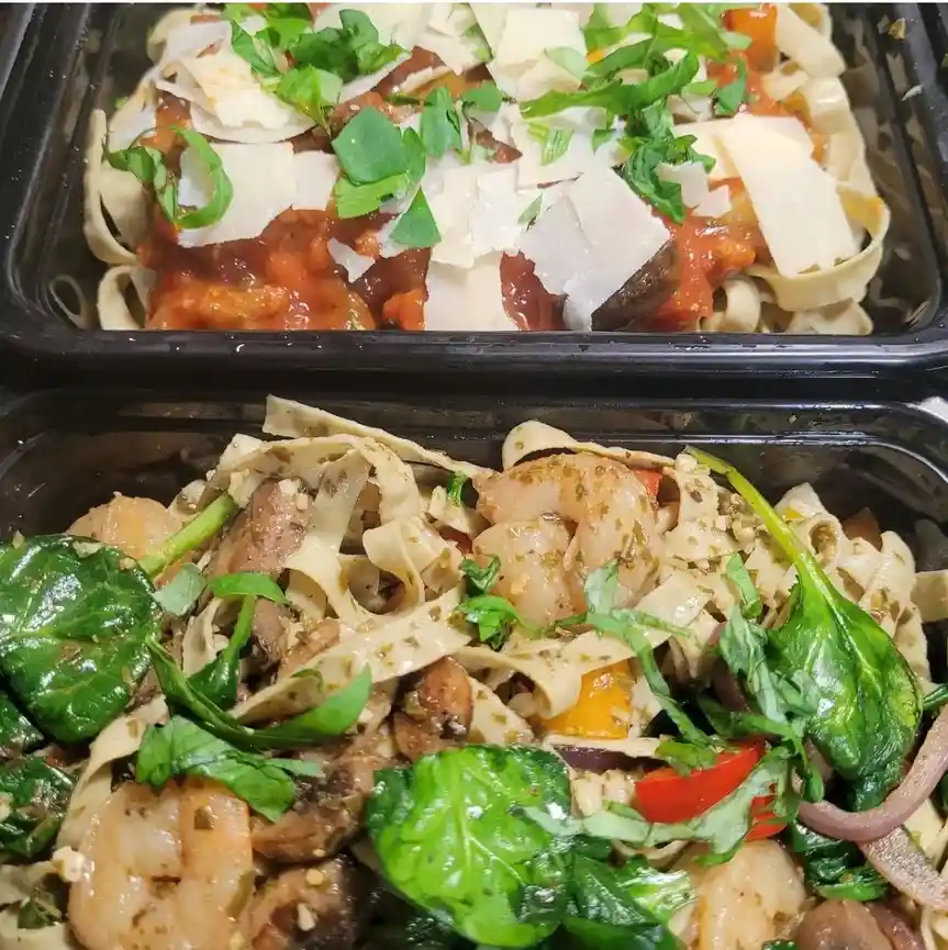 Two black takeout containers show pasta meals side-by-side. The top container holds pasta with red tomato sauce, topped with…