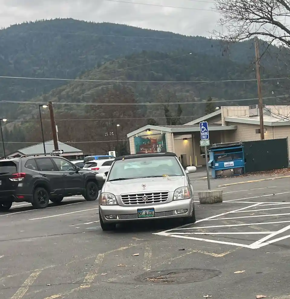 A silver Cadillac sedan is parked in a handicapped parking space marked with a blue and white wheelchair symbol sign. The…