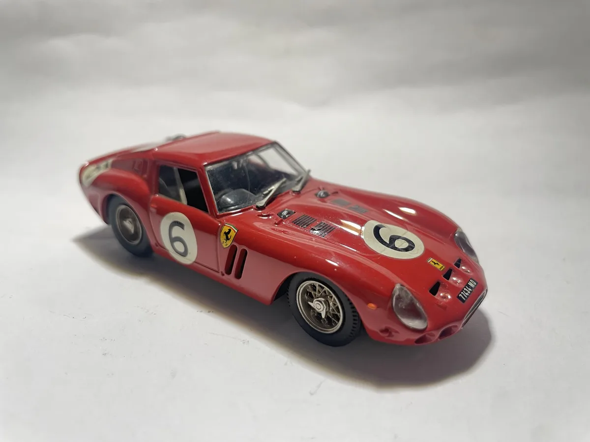 A red die-cast model of a classic Ferrari race car, featuring the number 6 on both sides, is photographed against a plain…