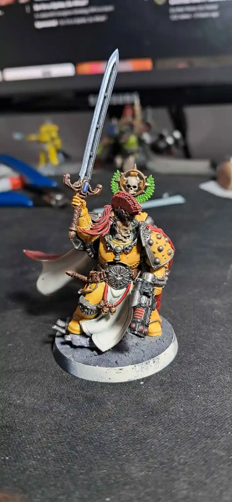 A heavily painted miniature of a grimdark fantasy warrior in yellow and silver armor, holding a large sword aloft while…