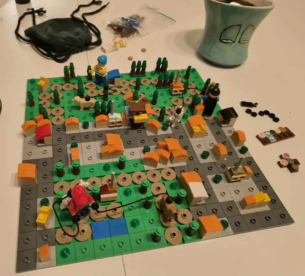 A tabletop scene shows a LEGO city built on green and gray baseplates, featuring LEGO minifigures, small houses, trees, and…