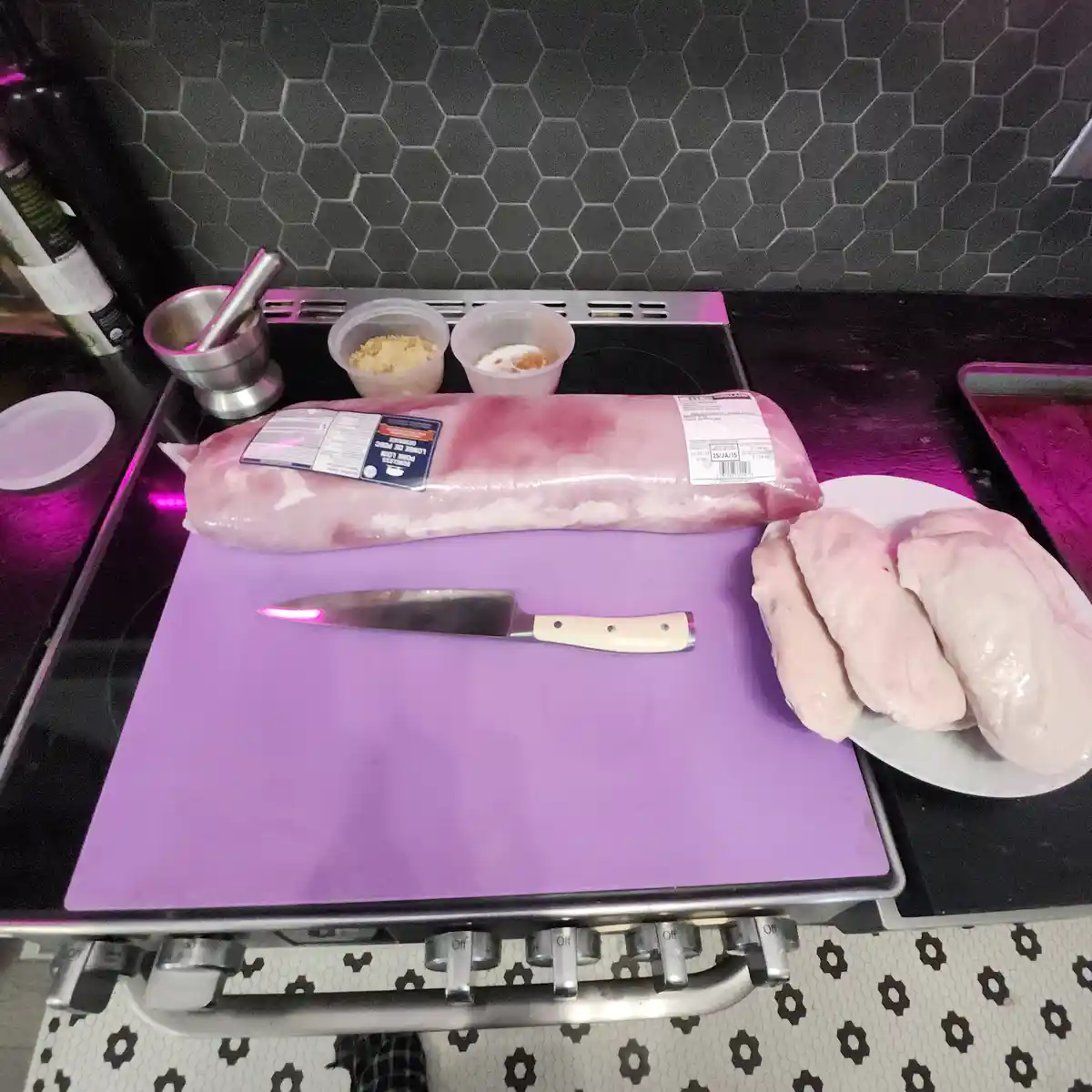 A kitchen scene shows raw pork loin on a purple cutting board next to a knife, with sliced chicken breasts on a plate…