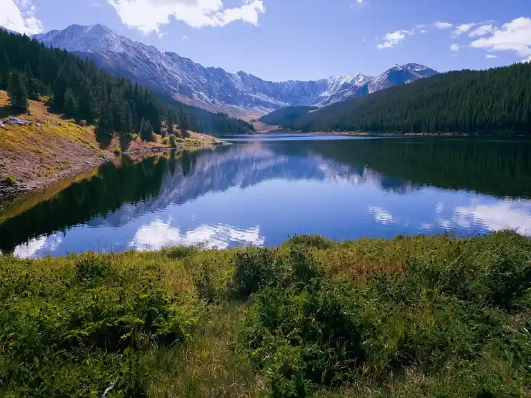 A serene mountain lake with calm, reflective waters mirroring snow-capped peaks and a blue sky with clouds. The foreground…