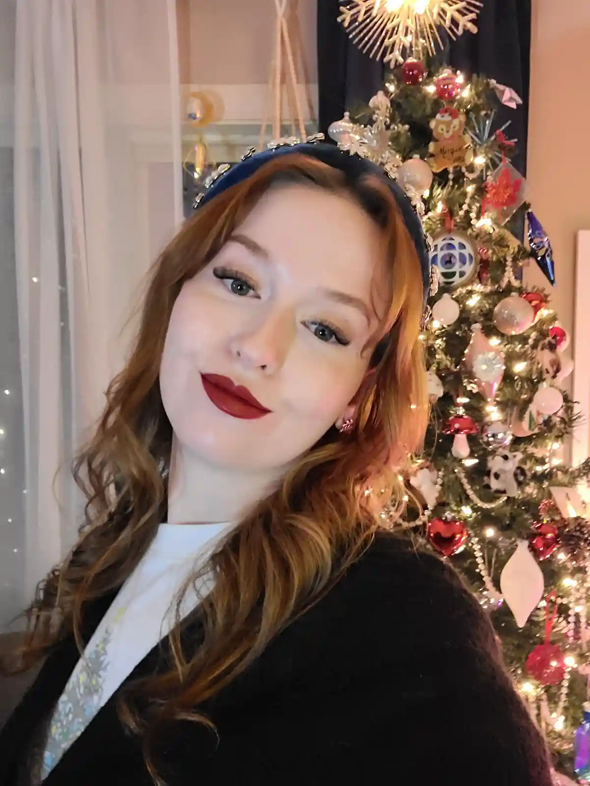 A woman with red hair and dark red lipstick smiles at the camera while wearing a blue velvet headband with silver…