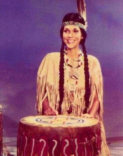 A woman with long braids and a feathered headband, wearing a fringed tan outfit and beaded necklaces, smiles while standing…