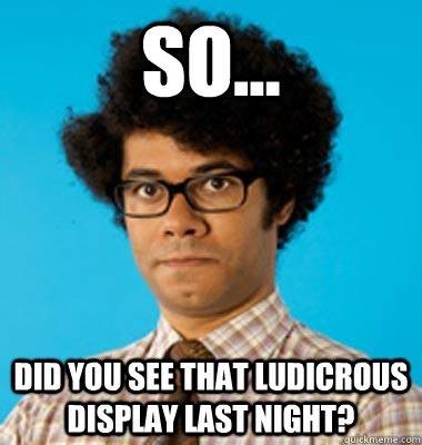 A man with a large afro, thick-rimmed glasses, and a plaid shirt stares blankly at the camera against a solid blue…