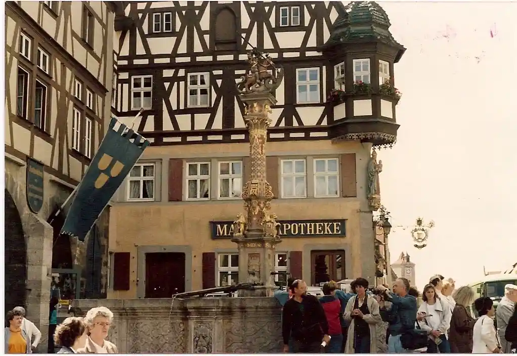 An old photograph shows a European town square with half-timbered buildings, a central ornate fountain, and tourists. A blue…