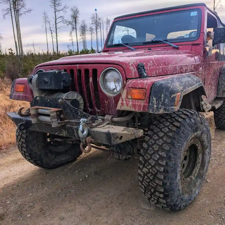 A red, mud-splattered Jeep Wrangler with large off-road tires and a heavy-duty front bumper sits on a dirt path in a wooded…