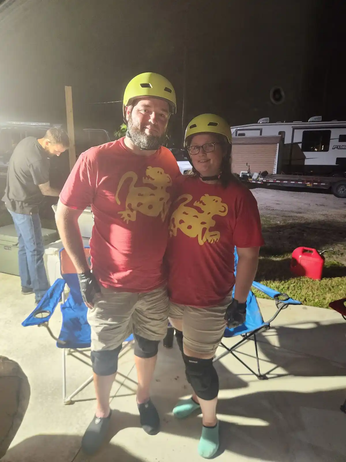 A man and woman wearing matching red t-shirts with a yellow lizard graphic and identical yellow helmets pose for a photo…