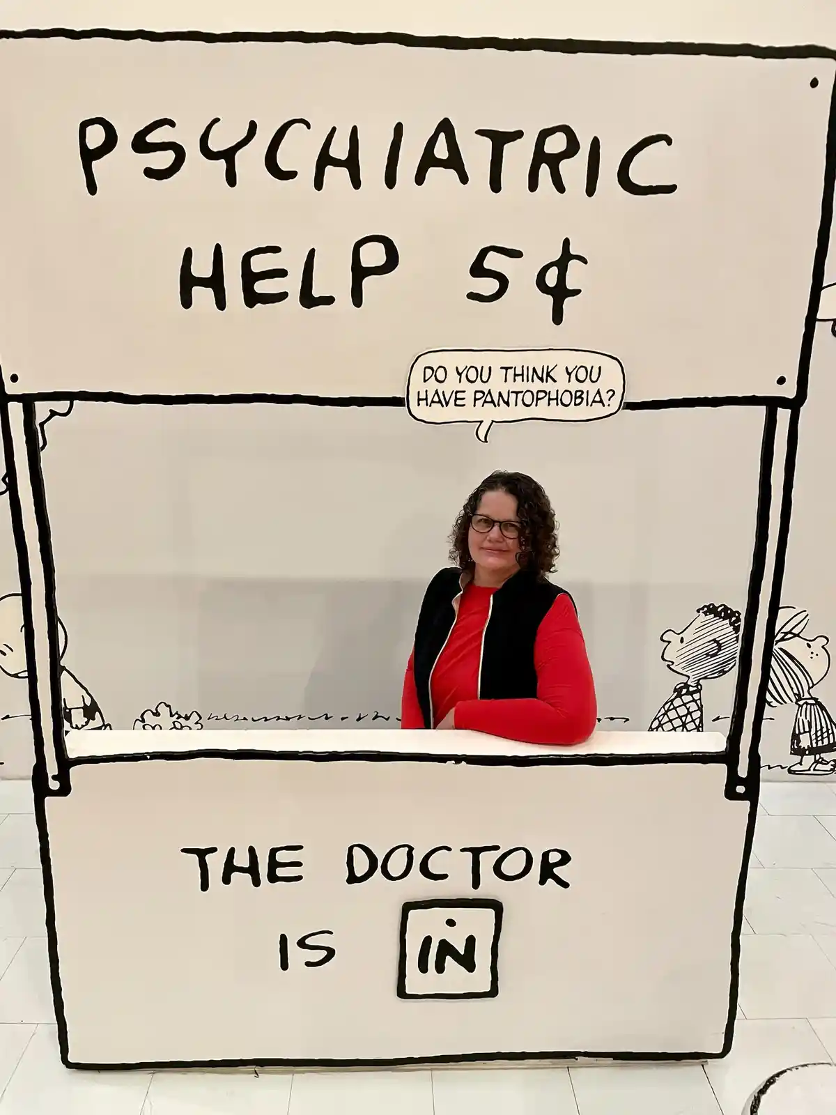 A woman with curly hair and glasses stands behind a white, hand-drawn booth resembling a psychiatrist's stand, labeled…