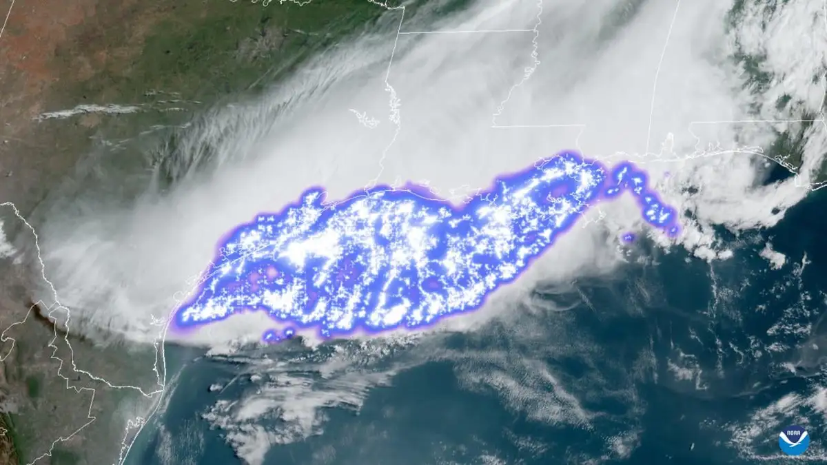 A satellite weather image shows the Gulf Coast region outlined in a glowing blue-purple aura, with white clouds and…