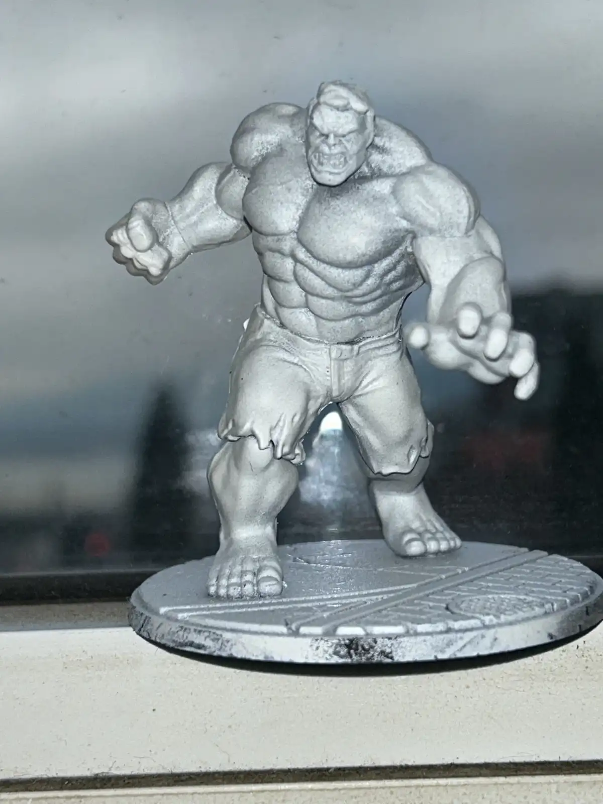 A gray, unpainted miniature figurine of the Hulk stands on a circular base, posed aggressively with clenched fists. The…