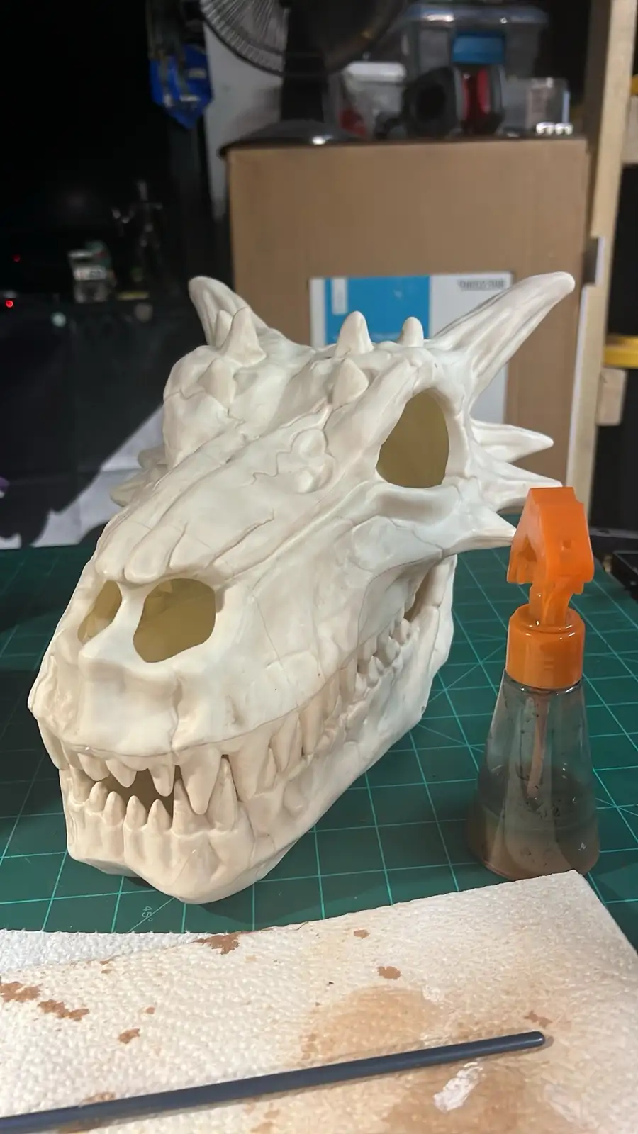 A large, white, unpainted dragon skull replica sits on a green cutting mat next to a small spray bottle with an orange…