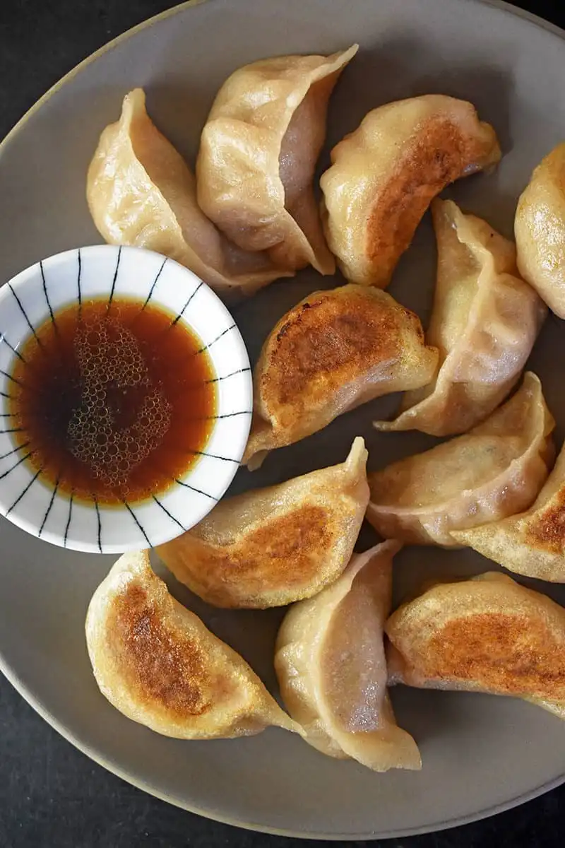 A top-down photo of a plate of golden-brown pan-fried dumplings with a small bowl of dipping sauce on the side. Overlaid…