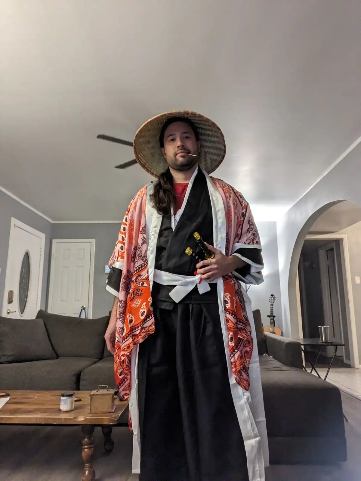 A man stands in a living room wearing a large straw hat, a black kimono with a colorful orange and pink patterned outer…