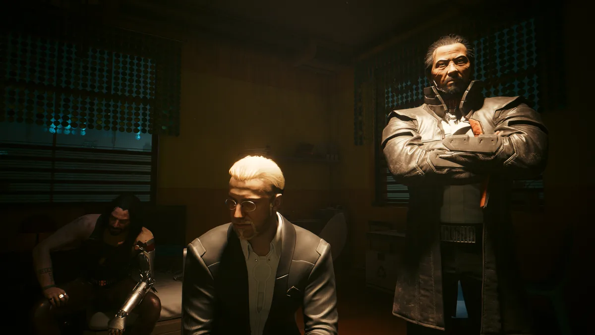 Three men are in a dimly lit room with perforated window blinds; one man in a suit sits center, another with a cybernetic…