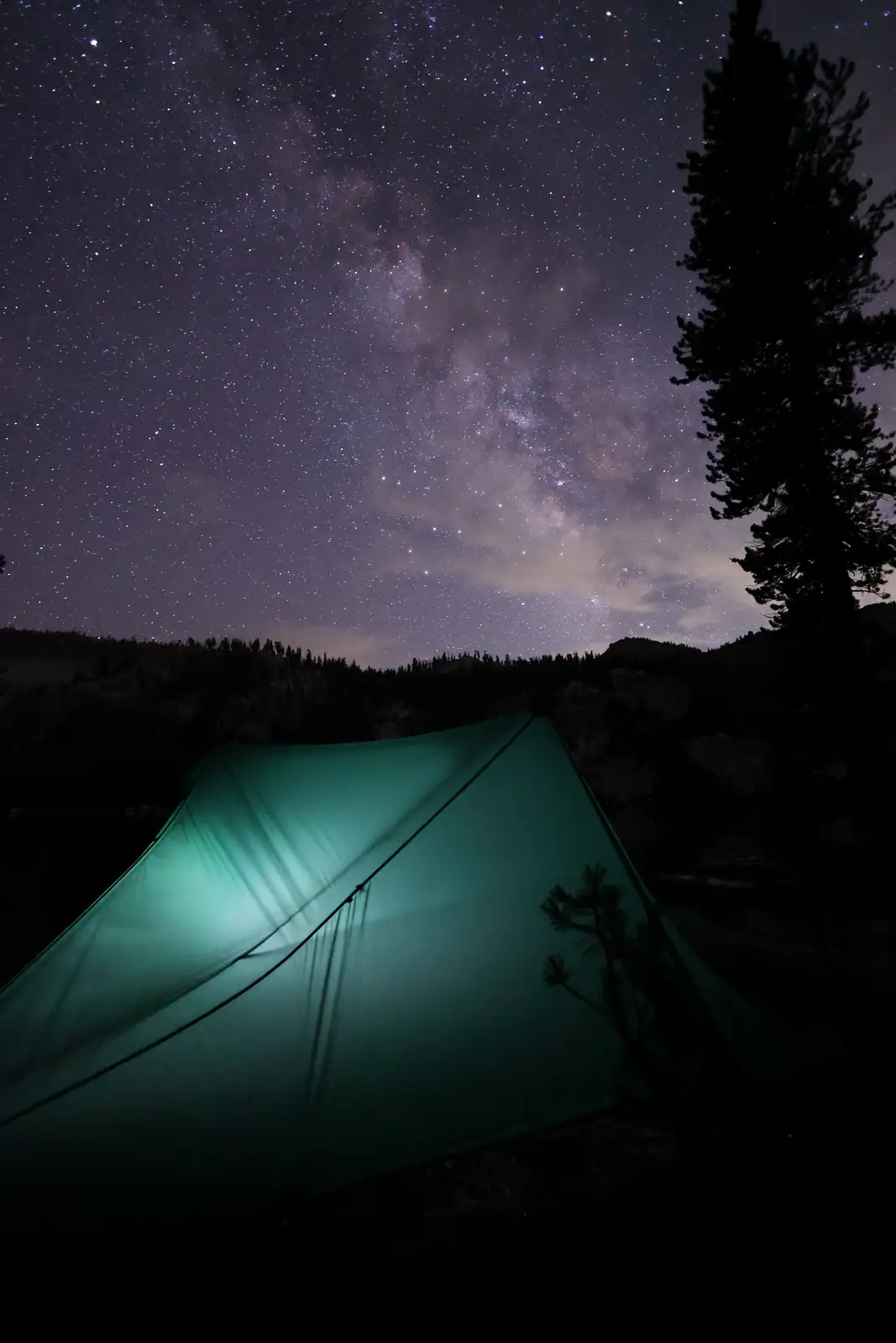 A green tent is illuminated from within, set against a dark landscape under a spectacular starry sky with the Milky Way…