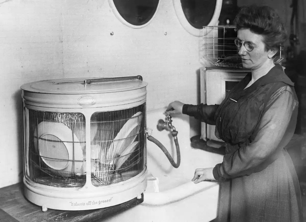 A black-and-white photo shows a woman in a vintage dress and glasses standing at a sink, turning on a faucet connected to an…