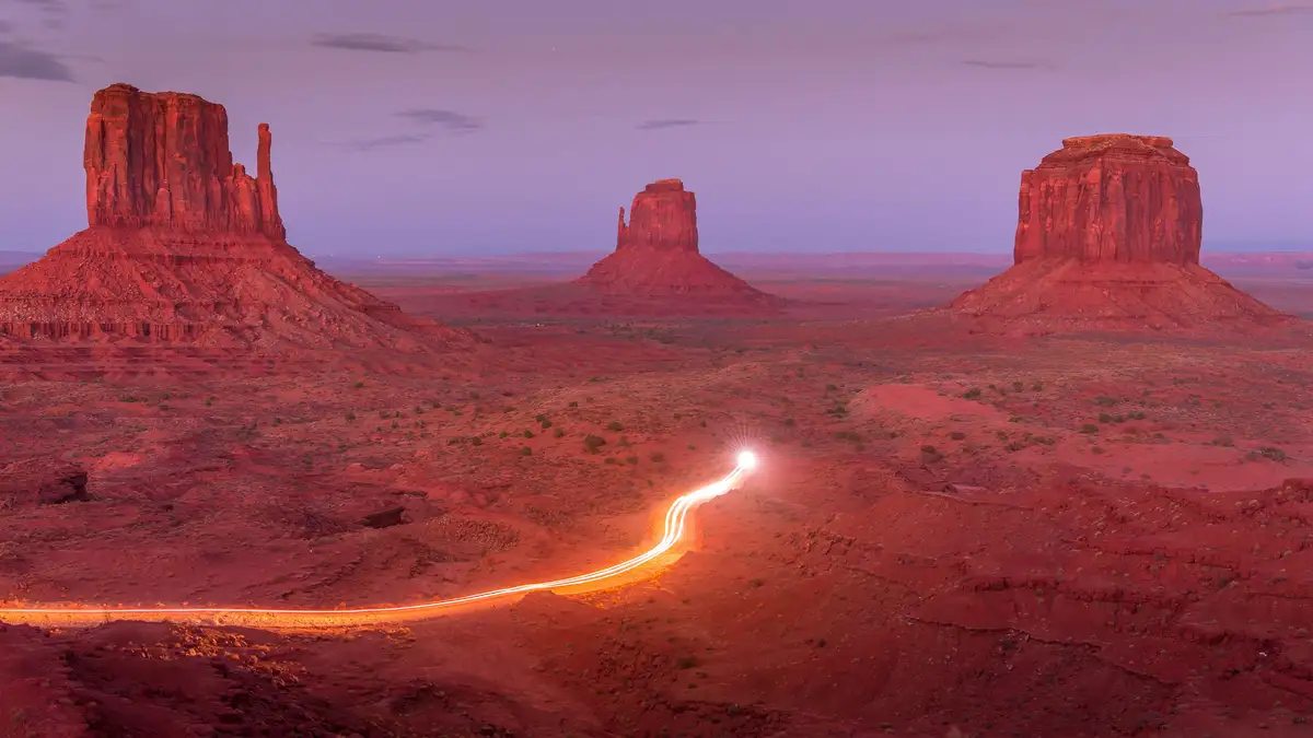Vibrant sandstone buttes ablaze with fiery hues, as the setting sun casts a warm glow on the rust-red landscape. Trails of…