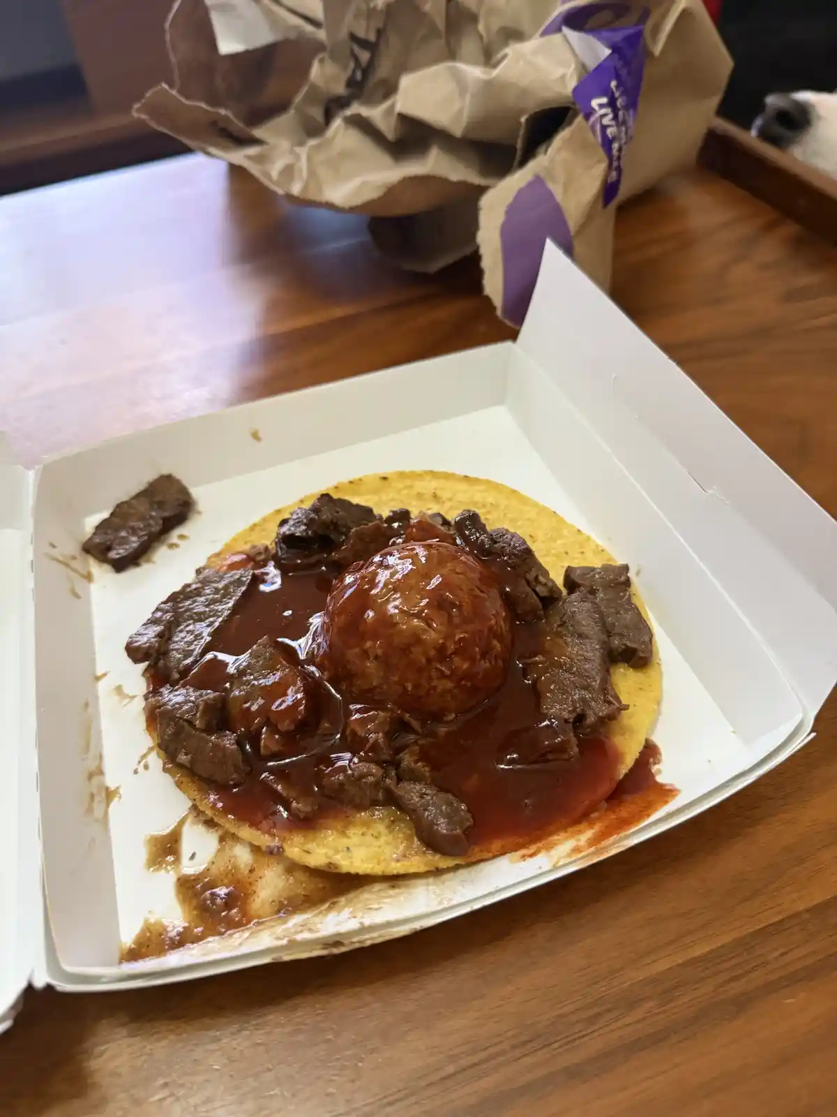 A close-up photo shows a taco in a white takeout box on a wooden table, featuring a large, round, glazed meatball centered…